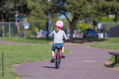Hillsboro, Oregon \ USA - 04 May 2019: A little girl in a pink helmet, blue capri and striped long sleeve shirt riding red bike on a school grounds