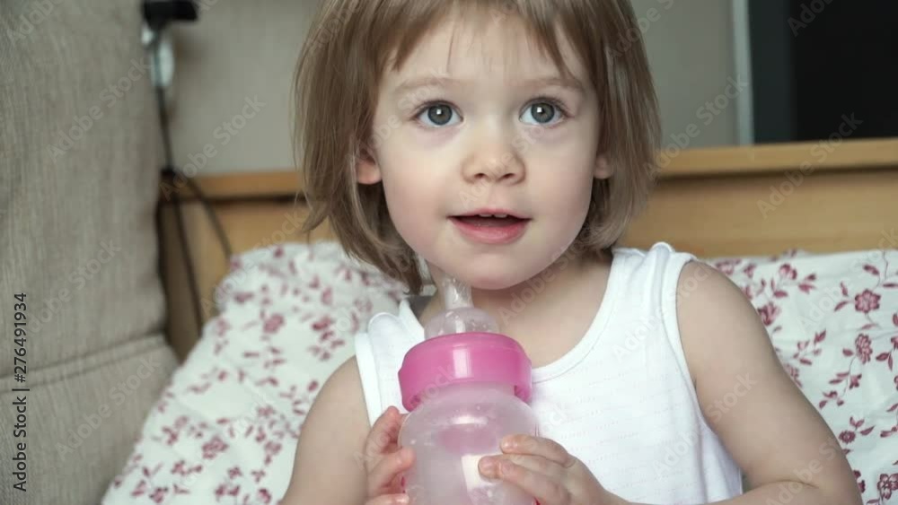 Portrait of little cute smiling baby girl sit on bed waking up in morning, holds in hands and hugs bottle of milk. Baby food formula concept Portrait of little cute smiling baby girl sit on bed waking up in morning, holds in hands and hugs bottle of milk. Baby food formula concept