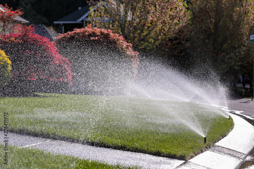 Water sprays from an automatic lawn sprinkler system over green lawn on a sunny day.