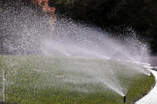 Water sprays from an automatic lawn sprinkler system over green lawn on a sunny day.