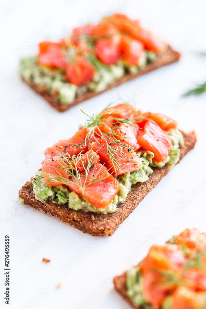 Close-up view of three sandwiches with rye bread, avocado and smoked salmon on a white kitchen table. Close-up view of three sandwiches with rye bread, avocado and smoked salmon on a white kitchen table.