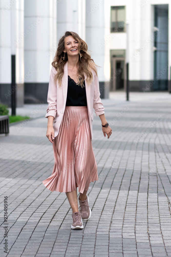 fashionable european girl walks down the street. Skirt plise, long jacket, sneakers. Beautiful long wavy hair. Peach, coral color clothes.