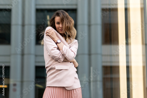 beautiful girl standing in a coral long pleated skirt, jacket, sneakers, black blouse. Wavy long hair hairstyle. Young european woman on the street near the glass building. Lifestyle summer.