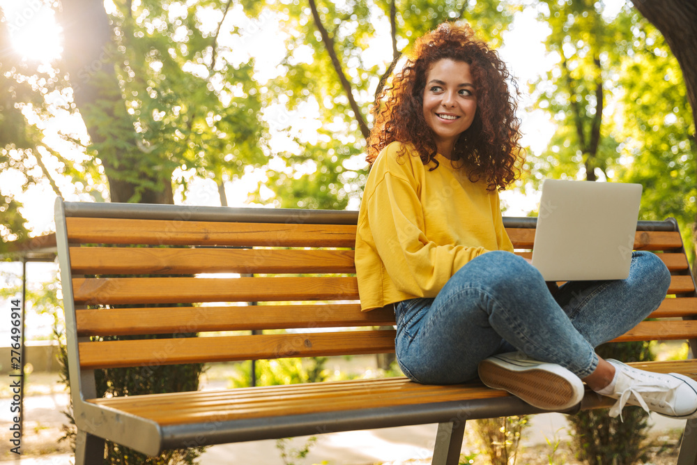 Happy young beautiful curly student girl sitting outdoors in nature park using laptop computer. - obrazy, fototapety, plakaty 