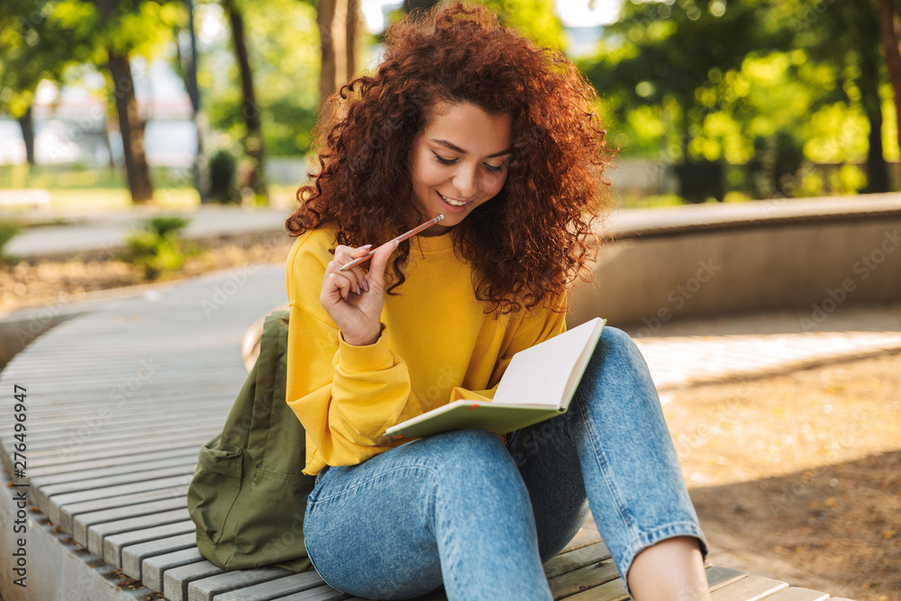 Young beautiful curly student girl sitting outdoors in nature park writing notes in notebook. - obrazy, fototapety, plakaty 