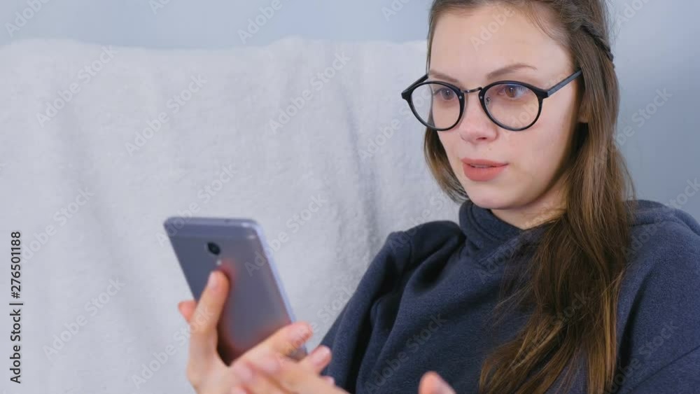 Happy young brunette woman in glasses talk a video chat on a mobile phone sitting in armchair Happy young brunette woman in glasses talk a video chat on a mobile phone sitting in armchair