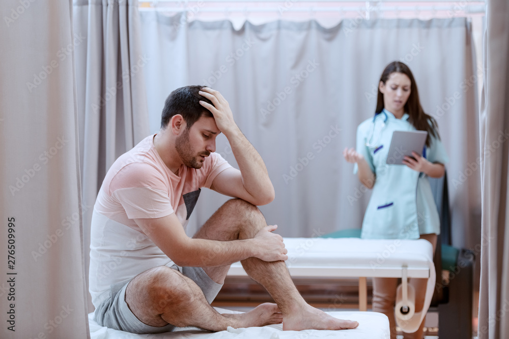 Young Caucasian male patient sitting on hospital bed and feeling pain. In background female nurse using tablet. Young Caucasian male patient sitting on hospital bed and feeling pain. In background female nurse using tablet.