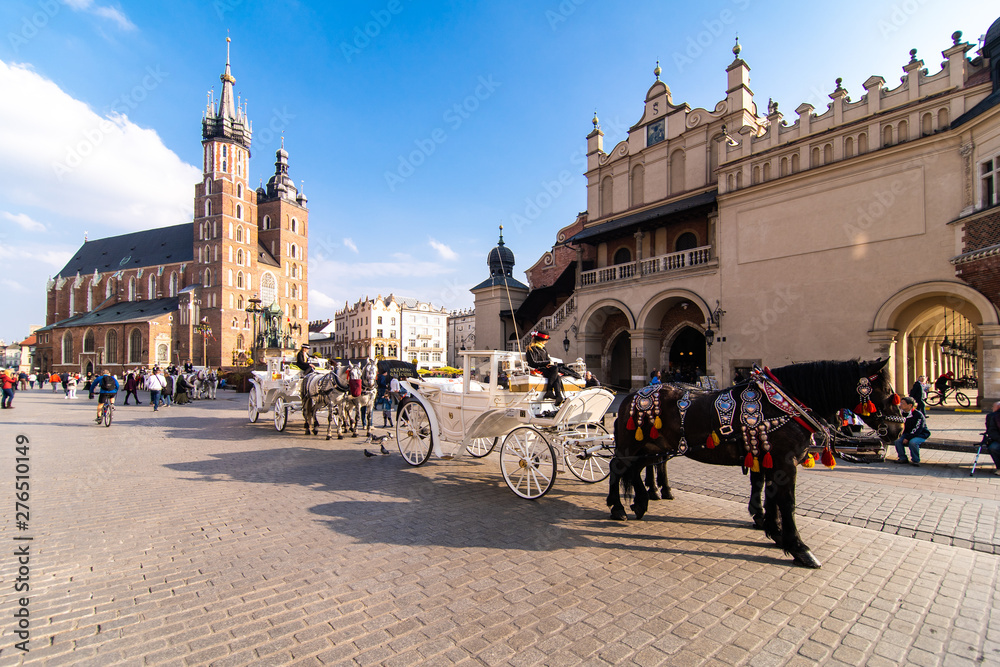 Obraz premium Kraków, Polska - kwiecień 2019: dwa konie w staromodnym powozie na Rynku Starego Miasta w pochmurny letni dzień. Bazylika Mariacka Słynny Punkt Orientacyjny Na Tle. Kościół Matki Bożej Wniebowziętej.