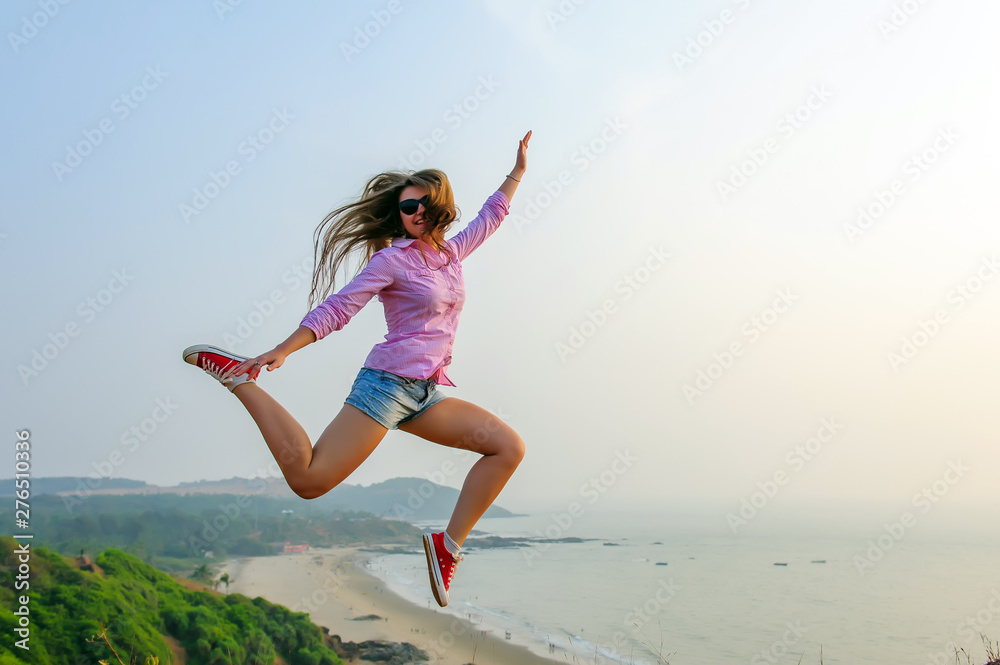 Young long-haired girl in shorts and red sneakers jumps high on the background of stunningly beautiful landscape with the sea and hills. Youth, freedom, travel, adventure. Young long-haired girl in shorts and red sneakers jumps high on the background of stunningly beautiful landscape with the sea and hills. Youth, freedom, travel, adventure.