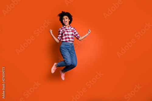 Full size photo of excited crazy jumping high lady yelling yeah raise fists wear jeans denim pants plaid shirt outfit isolated orange background © deagreez