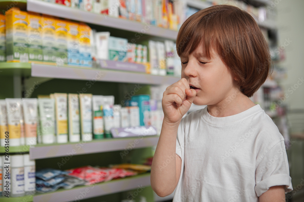 Close up of a little boy coughing, feeling sick at the drugstore, copy space. Little ill child coughing, covering his mouth with his hand