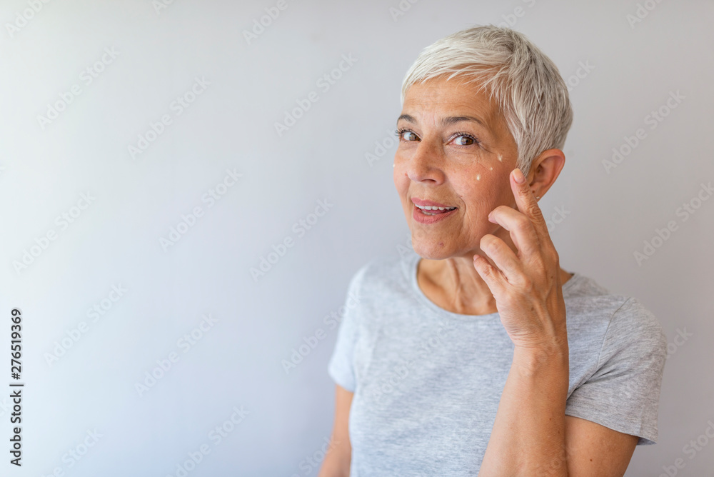 Smiling senior woman applying anti-aging lotion to remove dark circles under eyes. Happy mature woman using cosmetic cream to hide wrinkles below eyes. Lady using day moisturizer. Smiling senior woman applying anti-aging lotion to remove dark circles under eyes. Happy mature woman using cosmetic cream to hide wrinkles below eyes. Lady using day moisturizer.