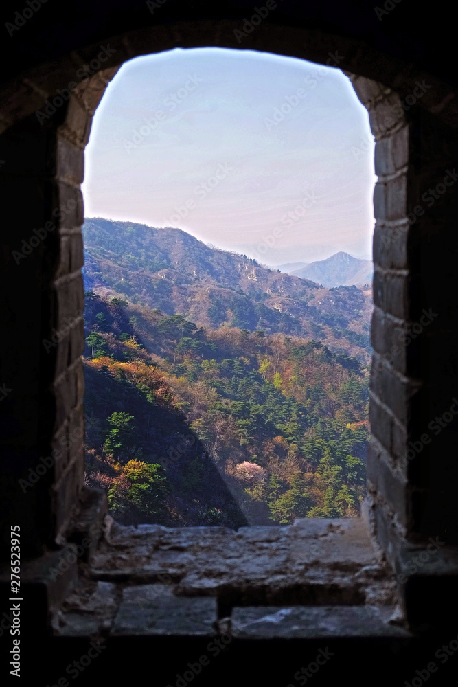 View from the window of a watch tower of the Mutianyu section of the Great Wall of China, surrounded by green and yellow vegetation under a blue sky.