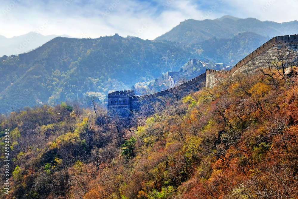 Panoramic view of the Mutianyu section of the Great Wall of China, surrounded by green and yellow vegetation under a cold morning light.