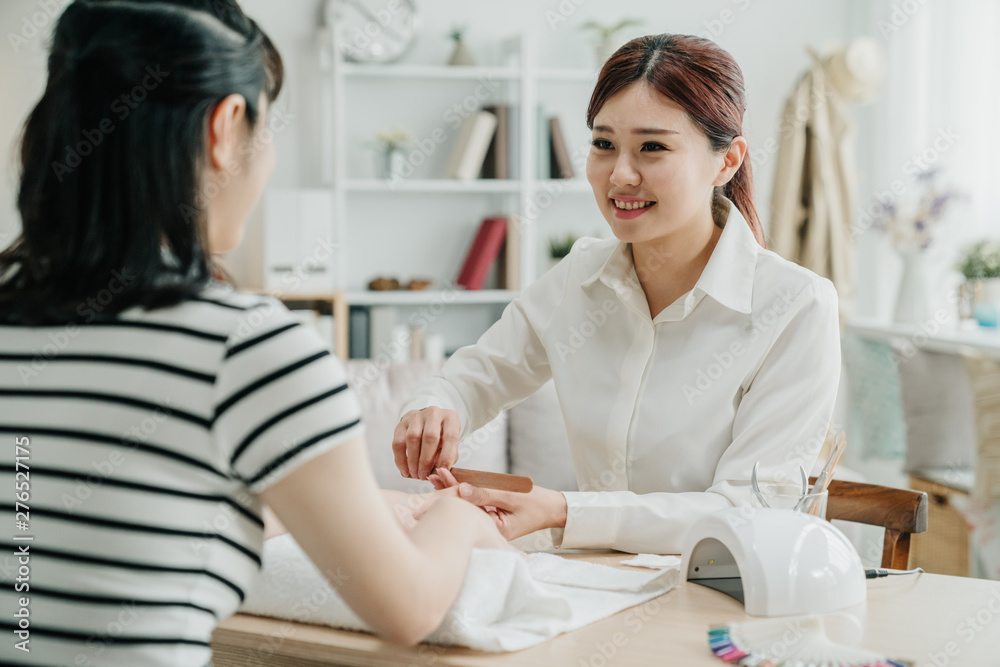 Obraz premium back view of female customer at home receiving manicure by smiling beautician with nail file. friendly manicurist file nails to lady client. elegant nail specialist working in guest house.