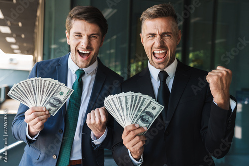 Image of excited businessmen partners rejoicing and holding bunch of cash money while standing outside office center