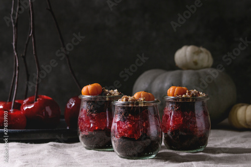 Layered Halloween dessert chocolate biscuit, raspberry jelly, nuts, marzipan pumpkin in glass jars and red caramel apples on branches served on linen table cloth with decorative pumpkins. Dark mood.