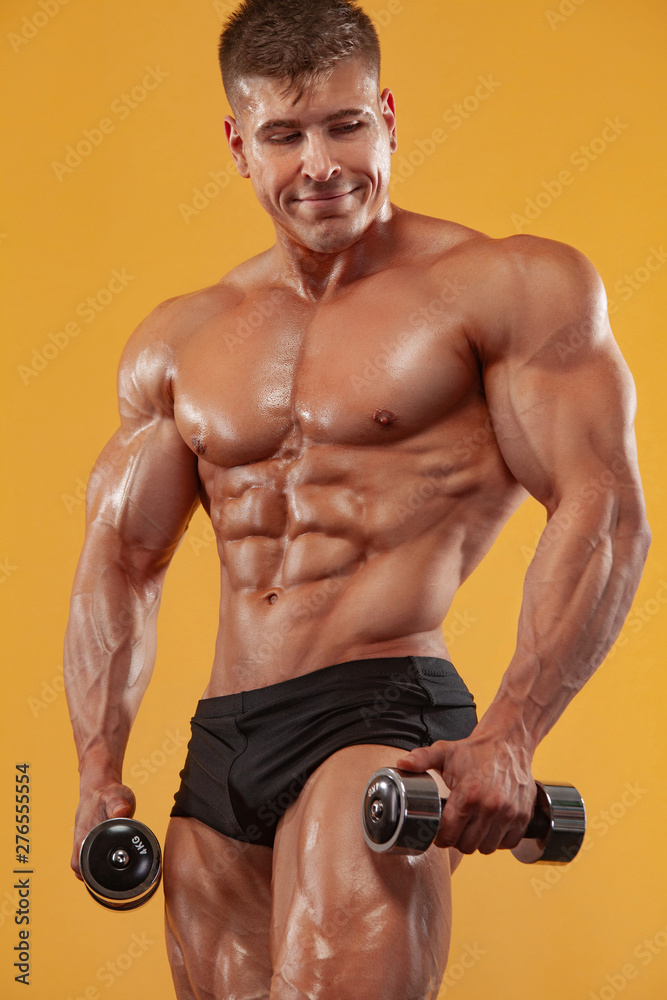 Brutal strong muscular bodybuilder athletic man pumping up muscles with dumbbell on yellow background. Workout bodybuilding concept.