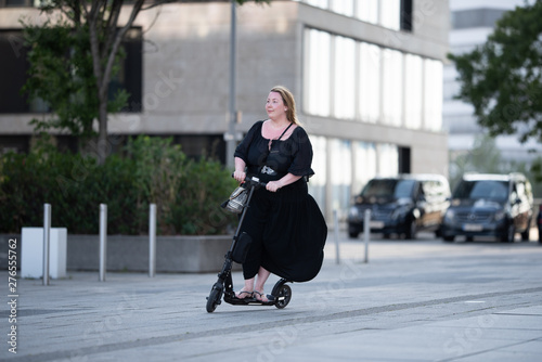 Portrait of a big business woman in black dress riding an electric scooter in urban environment next to parking cars at media harbor duesseldorf after work
