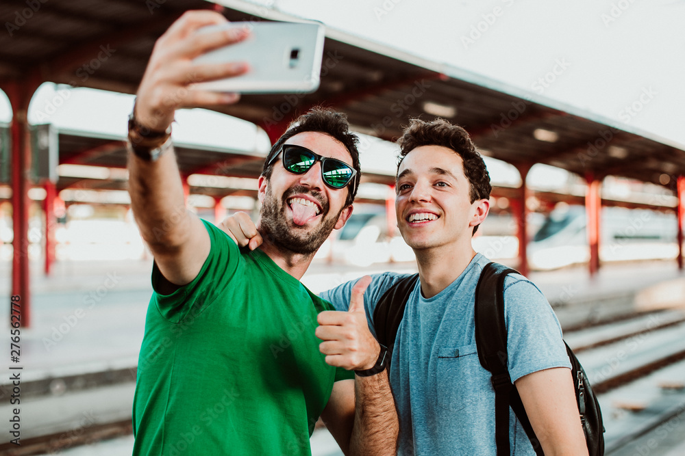 .Two brothers on the platform waiting for the train to start their summer vacation. Taking pictures together with their cellphone. Lifestyle. Travel photography - obrazy, fototapety, plakaty 
