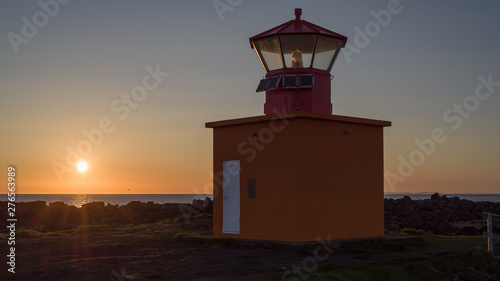 Sunset on west coast of Iceland - low sun above Atlantic with a lighthouse in foreground. Back light (close to midnight) throws beams on a rocky coastline dominated by a red land orange lighthouse.