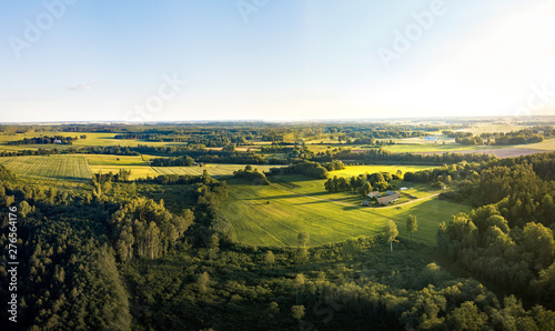 Fototapeta Naklejka Na Ścianę i Meble -  Aerial view over rural landscape in a warm summer sunset tones. Agriculture land mixing with forest and meadows. Green crop fields along the curved river. Trees creating long shadows. 