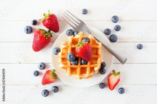 Breakfast waffles with scattered strawberries and blueberries. Top view over a white wood background.