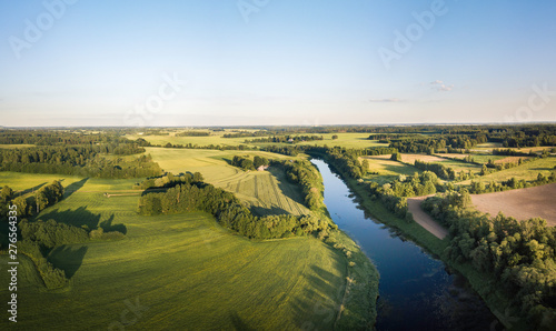 Aerial view over rural landscape in a warm summer sunset tones. Agriculture land mixing with forest and meadows. Green crop fields along the curved river. Trees creating long shadows. © Viesturs