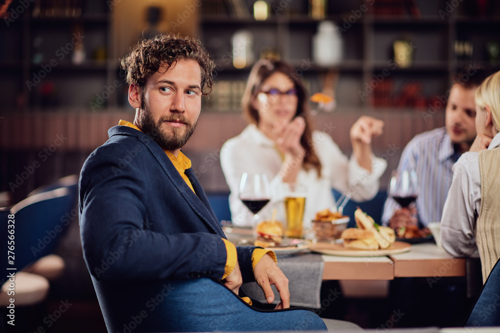 Young serious man dressed smart casual sitting at restaurant and looking over shoulder. In background his friends having dinner.