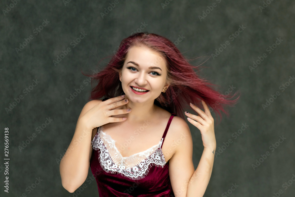 Concept portrait of a pretty sexy girl with purple hair in beautiful red lingerie on a gray background. She smiles, happy with life, happy. Hair flying, front view. Made in a studio.