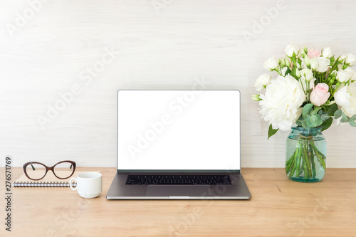Office desk table with laptop computer with blank screen mockup and flowers bouquet, coffee cup, glasses, notepad on wooden surface. Minimal style feminine workplace concept.