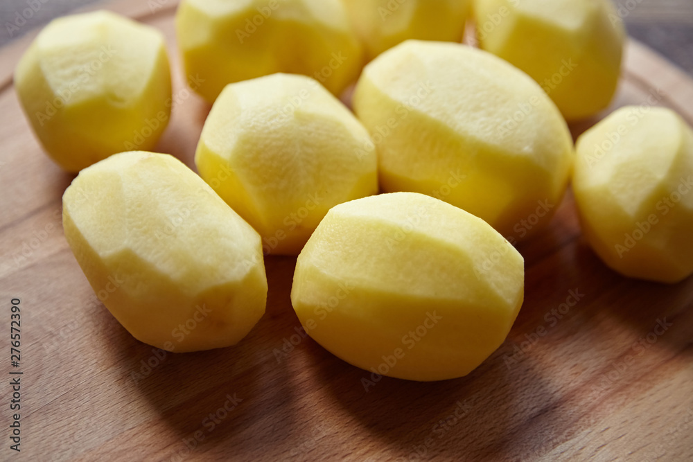 Peeled potatoes on wooden cutting board on brown table. Cooking food from natural products. Root vegetable. Raw ingredient: uncooked whole potatoes