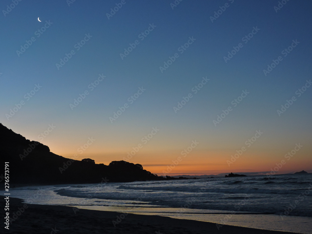 Atardecer con cielo azul y naranja, un monte y la luna en la playa