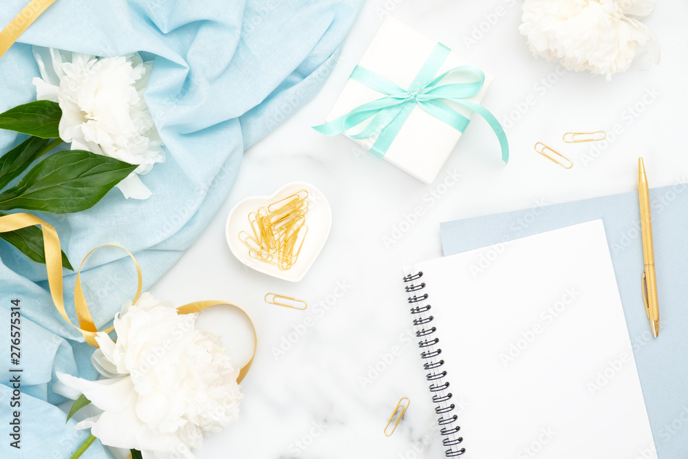Top view wedding woman accessories on marble background. Minimal flat lay style composition with golden clips, white peony flowers, blank paper notebook, gift box. Wedding and marriage concept.