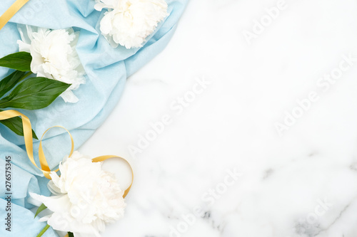 Top view white peony flowers, blue scarf and golden ribbon on marble background. Minimal flat lay style home desk with peonies Wedding invitation card mockup. Beauty or fashion blog banner template