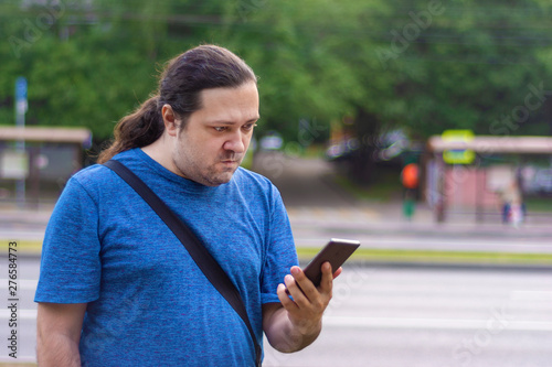 A middle-aged man in casual clothes angrily looks at the phone screen on the street on a summer day.