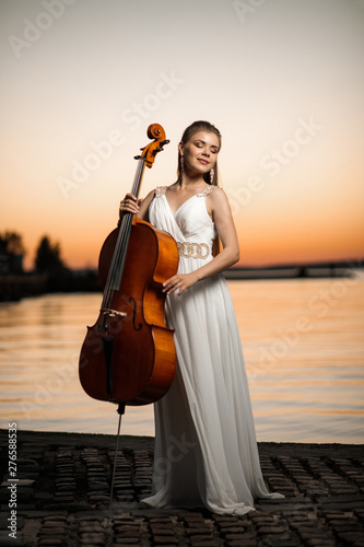 Stampa su tela girl in white beautiful dress with a cello stands by the lake at sunset
