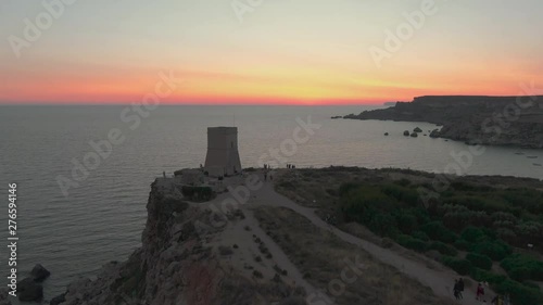 aerial drone dolly in shot approaching Għajn Tuffieħa Tower (Torri t'Ghajn Tuffieha), on golden sunset. Tourists exploring Malta historical landmark and enjoying sunset photo