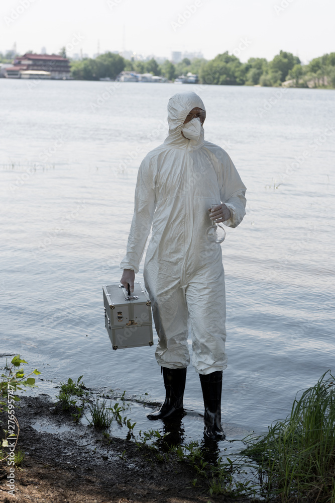full length view of water inspector in protective costume holding inspection kit and flask at river full length view of water inspector in protective costume holding inspection kit and flask at river