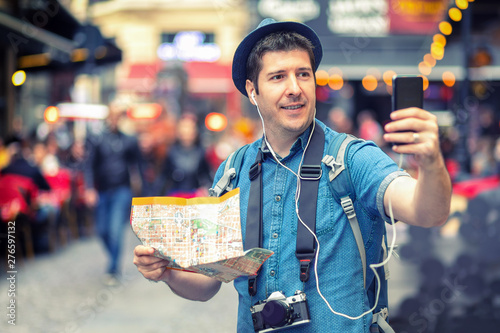 Smiling man tourist holding city map having roaming video call on mobile phone on European crowded streets full of pubs 