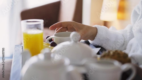 Girl enjoying morning in light hotel apartment with full length windows. Woman in white bathrobe having breakfast in bed, detail view tray full of food and female hand taking cup of tea 