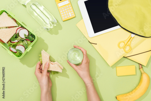 cropped view of woman hands with sandwich and glass of water near lunch box, backpack, digital tablet, bottle of water and stationery
