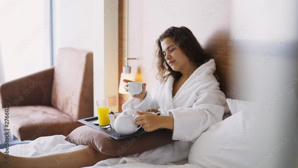 Girl in light hotel apartment with full length windows. View through glass woman in white bathrobe relaxing in bed and having breakfast on small table-tray, enjoying morning coffee Girl in light hotel apartment with full length windows. View through glass woman in white bathrobe relaxing in bed and having breakfast on small table-tray, enjoying morning coffee