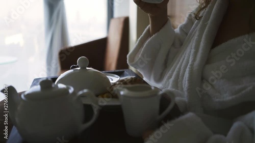 Woman have breakfast in modern hotel apartment with full length window. Close-up view food and pot on tray, girl taking cup of coffee 