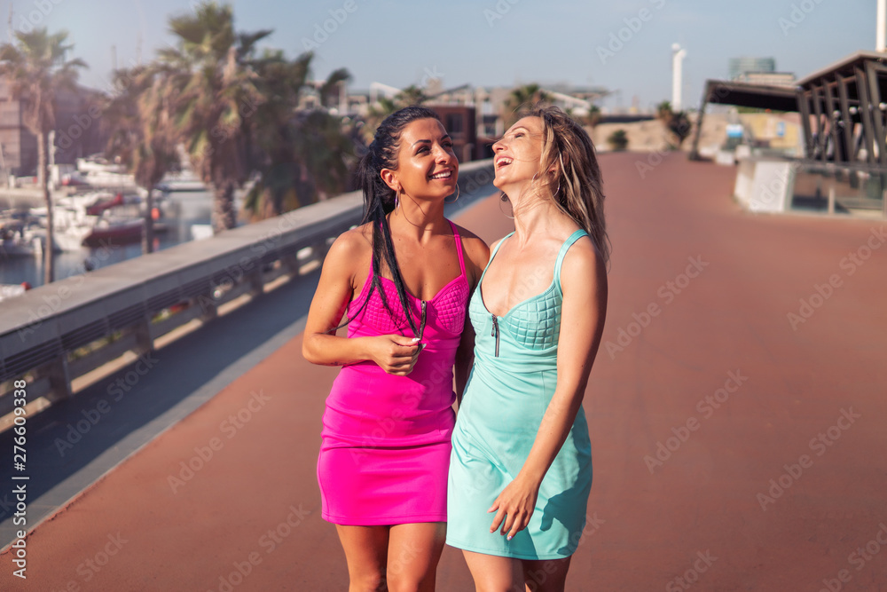 Happy best friends or sisters walking in the park with palm trees and yachts on background. They are posing and showing lovely relationship. They look happy.