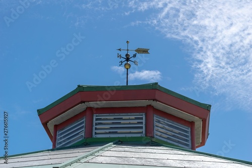 A weather vane atop an old building, against a blue sky with some light fluffy clouds