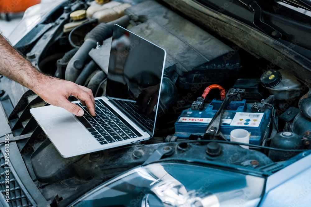 cropped view of car mechanic using laptop with blank screen near car