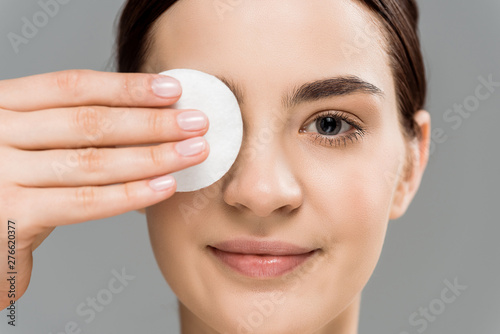 close up of cheerful naked woman covering eye with cotton pad isolated on grey