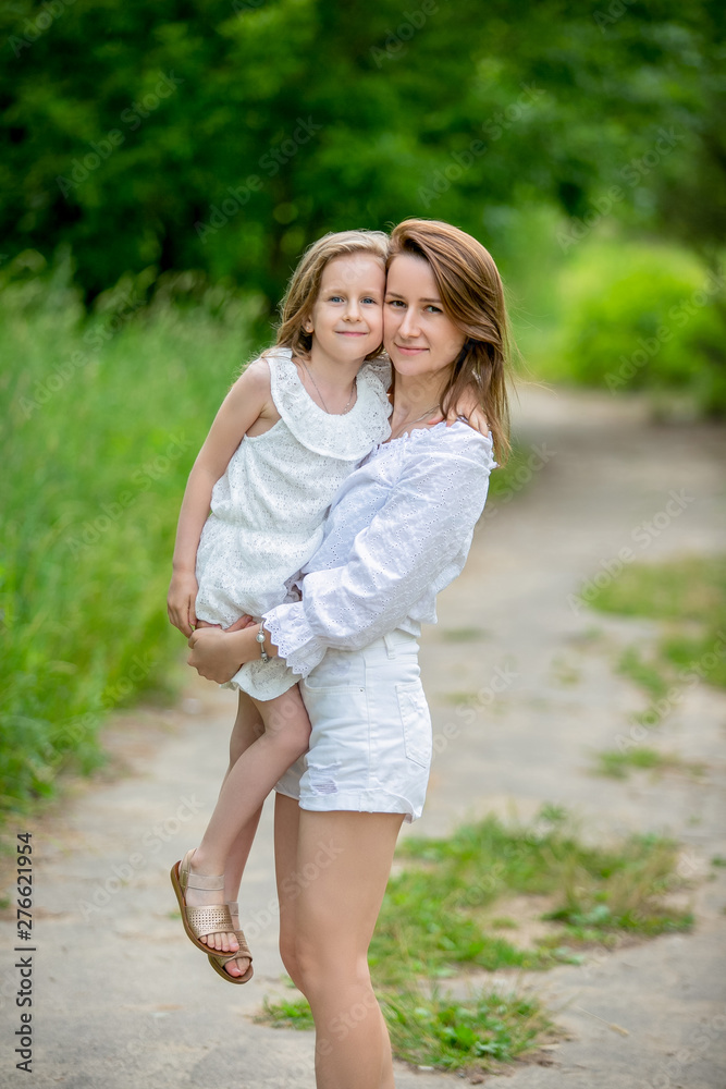 Beautiful young mother and her little daughter in white dress having fun in a picnic. They stand on a road in the park, mom holds her daughter in her arms. Maternal care and love Beautiful young mother and her little daughter in white dress having fun in a picnic. They stand on a road in the park, mom holds her daughter in her arms. Maternal care and love