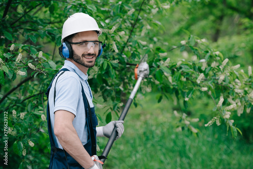 handsome gardener in helmet, protective glasses and noise-canceling headphones holding telescopic pole saw and smiling at camera